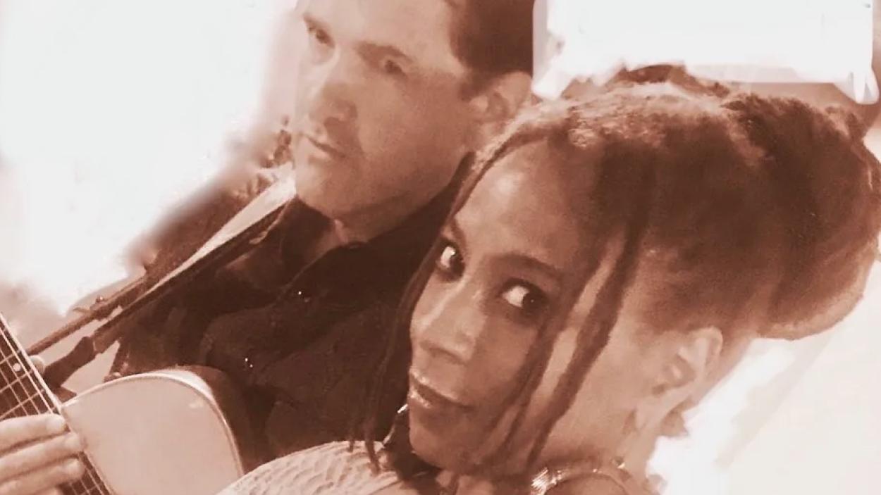 A brownish red tinted image of a light skinned man holding a guitar, and a dark skinned woman, tilted at an angle with a white background