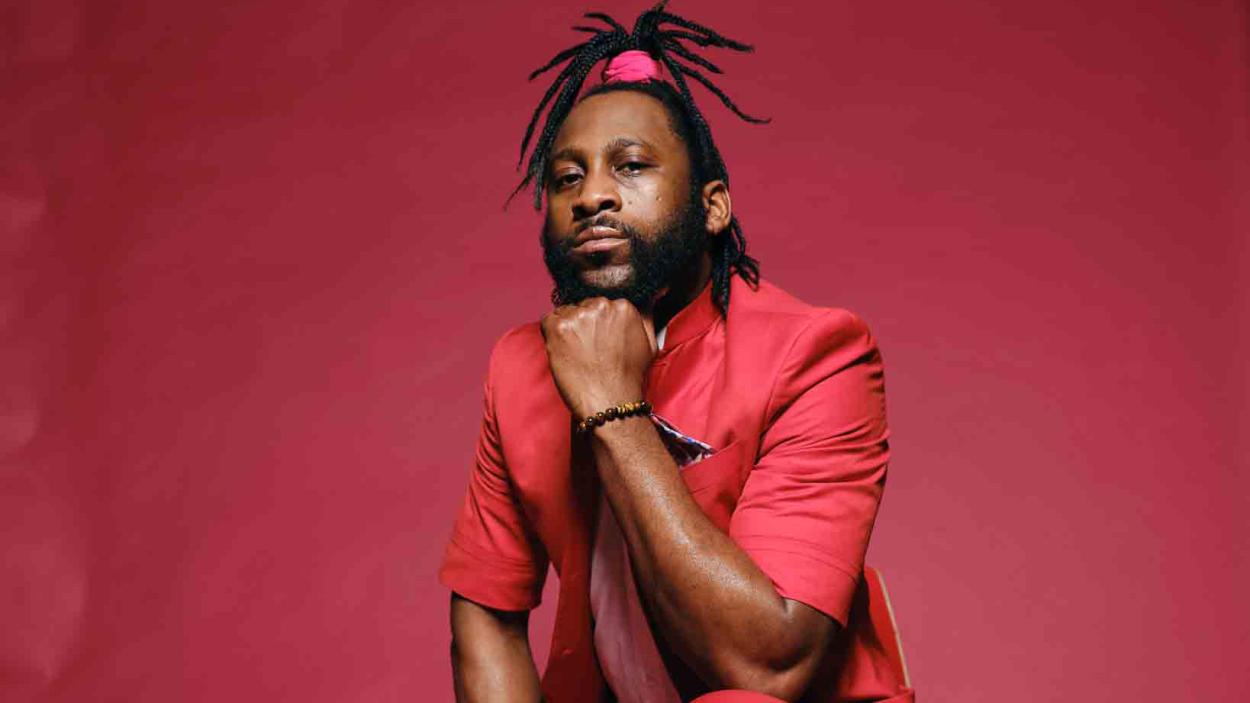 A dark skinned man wearing a red blazer, and dreadlocks in a top knot secured with a red band, sits in front of a red background, resting his chin on his hand