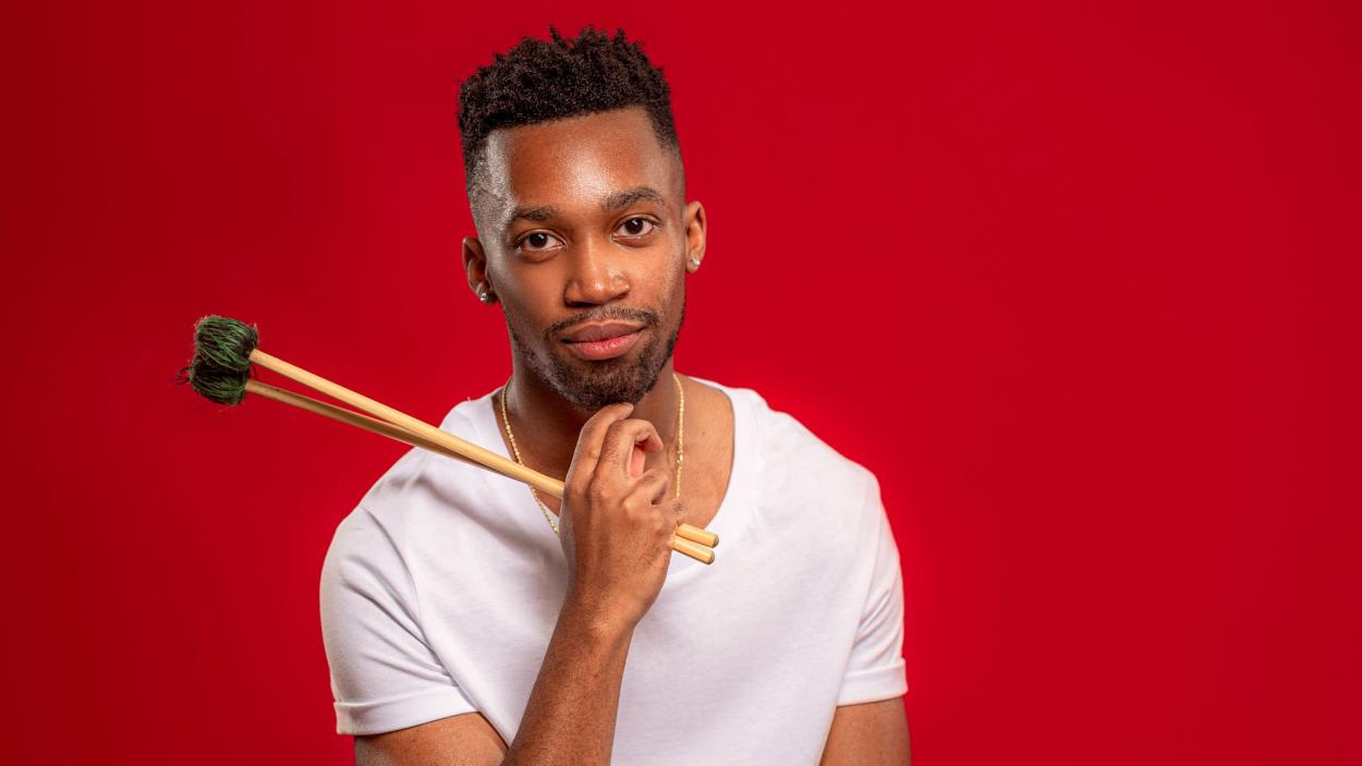 A dark skinned man sits in front of a red background, wearing a white tee shirt hold vibraphone sticks