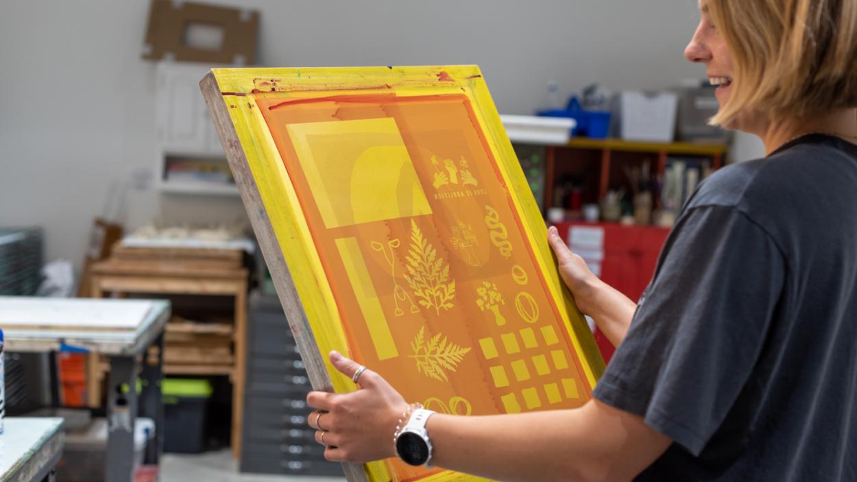 A light skinned woman with short blond har wearing a grey t-shirt holds a yellow and orange screen print with leaf shapes 