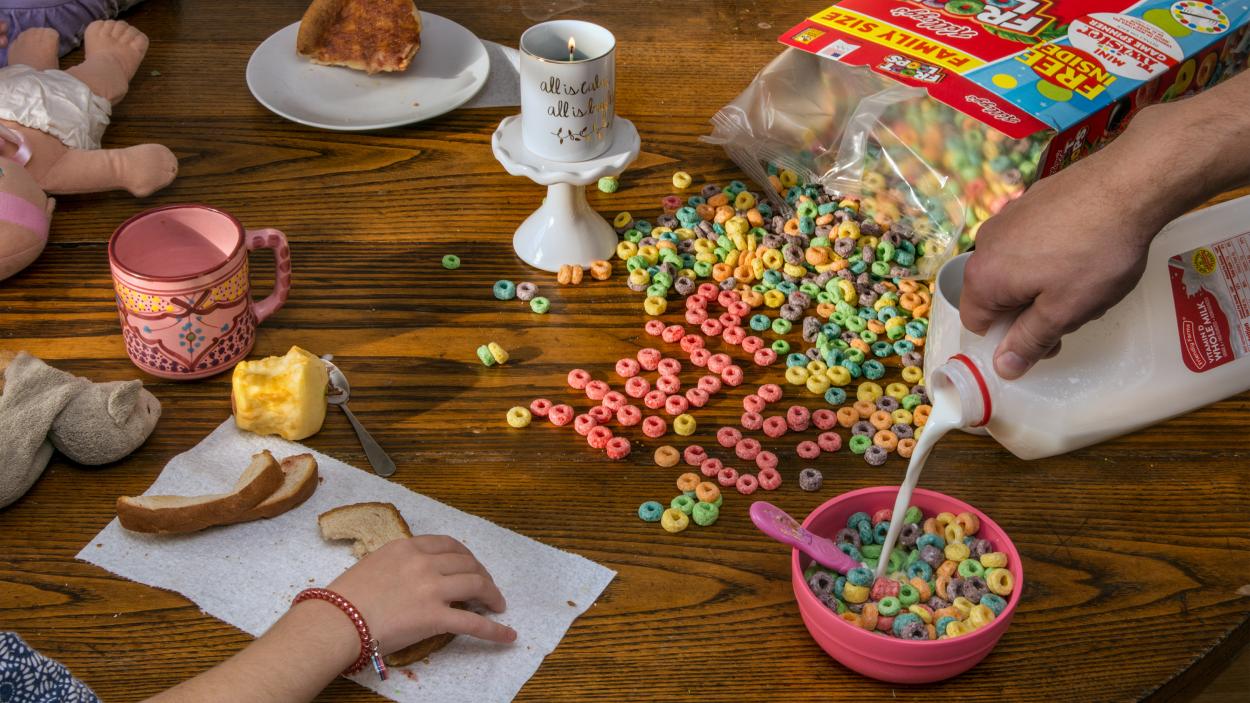 A photograph of a messy breakfast table. A box of fruit loops spills across the brown wooden surfac, with Help Us spelled out in pieces of cereal, while a hand reaches in to pour milk into a bowl of cereal. A childs hand grabs a half eaten piece of toast from a paper towel, and a mostly eaten apple sits on the table along with babydoll lying facedown a pink mug, a porcelain tea cup on a small pedestal and toast crusts.