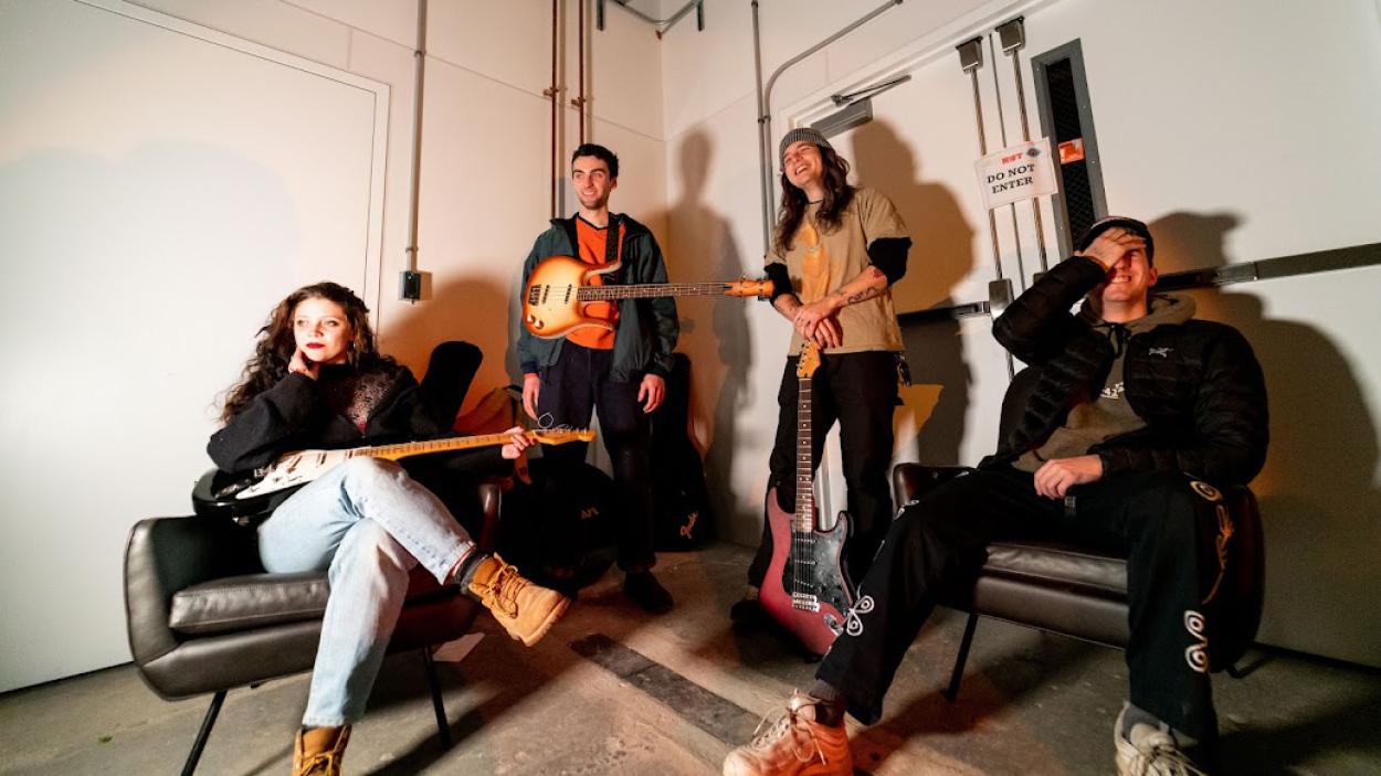 A light skinned woman with wavy brown hair and three light skinned men sit together in an industrial space with white walls