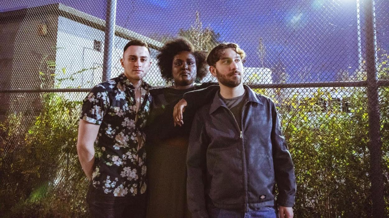 Two light skinned men and a dark skinned woman stand close together in front of a tall chain link fence at twilight, with green grass and a darkening sky behind