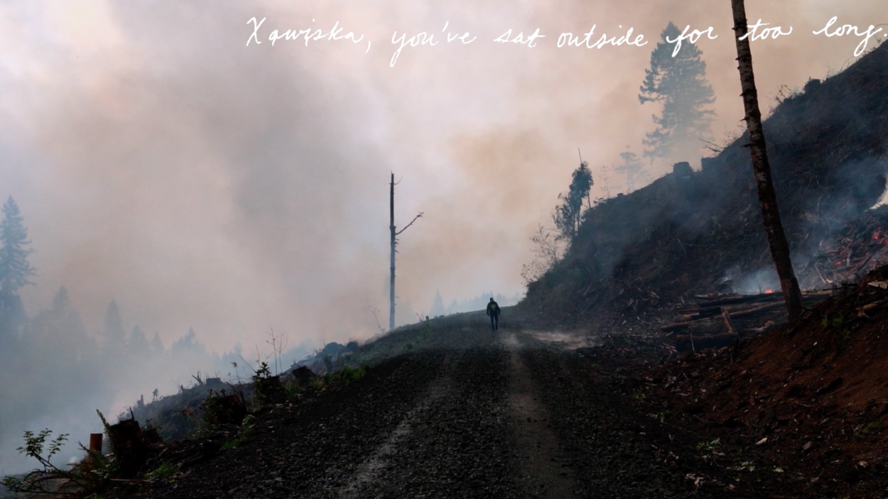 a still from a film that shows a man walking down a dirt road amidst a smoking landscape that has recently been burned with white handwriting that says " Xwįska, you've sat outside too long"