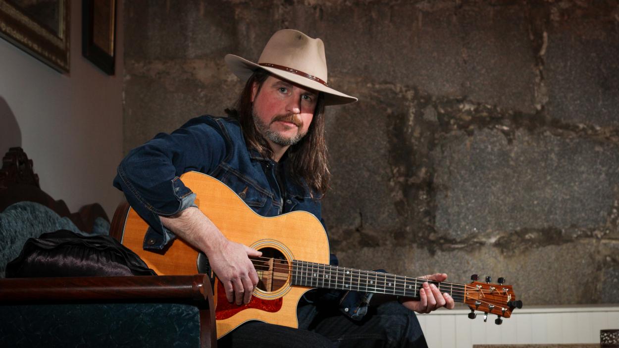 A light skinned man wearing a beige cowboy hat sits holding a honey colored acoustic guitar in front of a cracked concrete wall