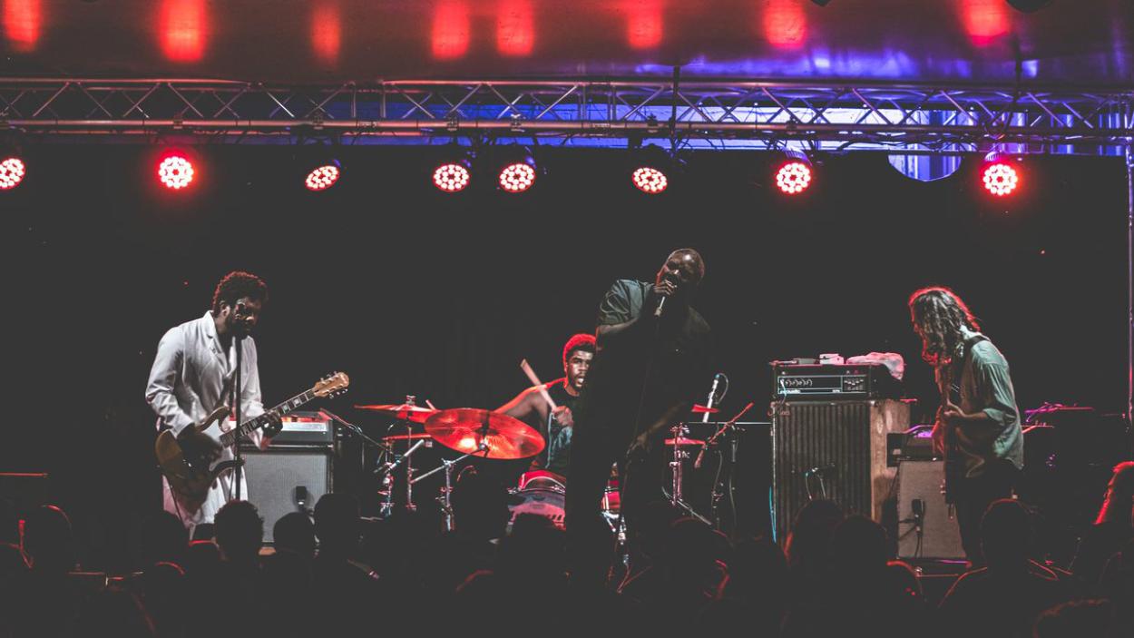 66b3d176-b8a8-4126-968f-378cdc52a183.jpeg A dark skinned man sings into a microphone, accompanied by a dark skinned man wearing a white shirt playing bass, a dark skinned man playing the drums and a light skinned man playing a guitar. Red and blue stage lights hang above them.