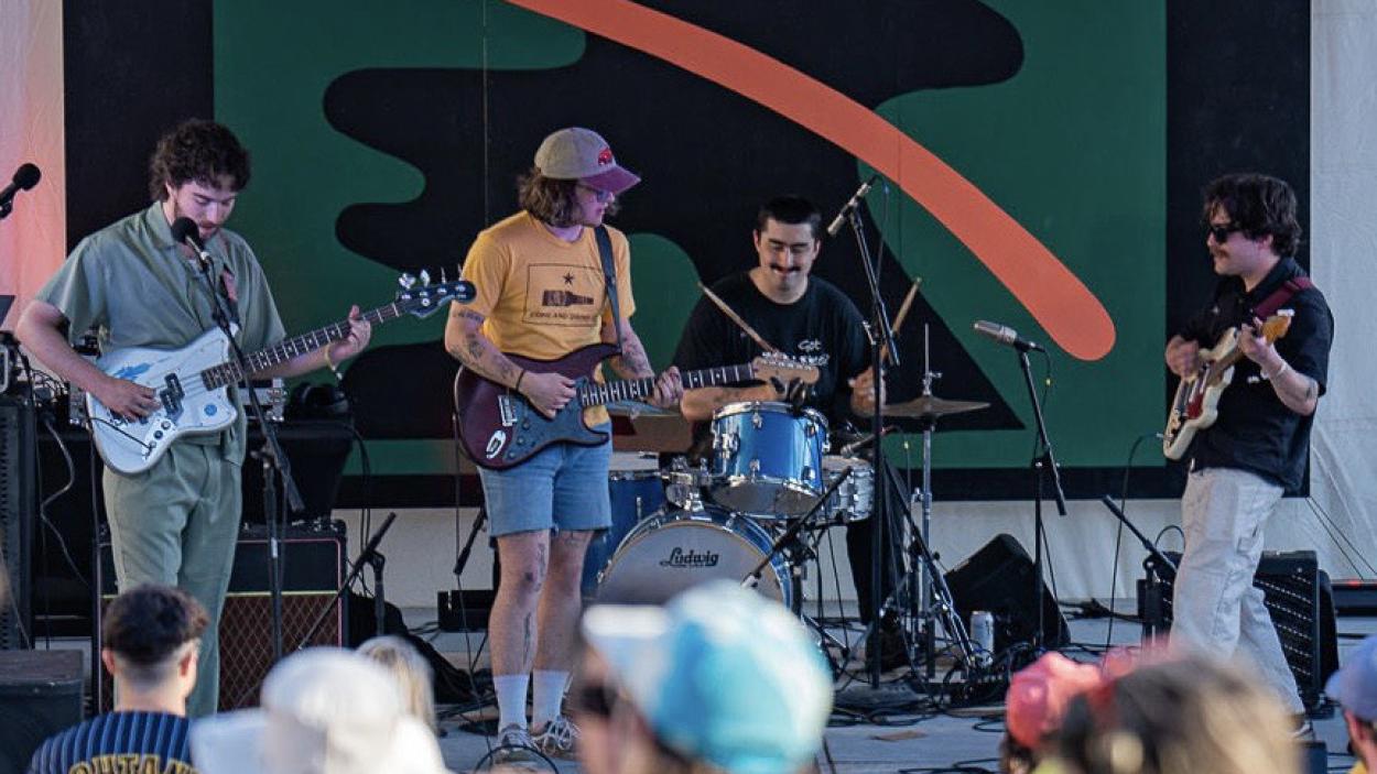 image002_1.jpg A band consisting of three light skinned men playing guitar and bass and one man playing drums stands on a stage with an abstract green and orange mural in the background