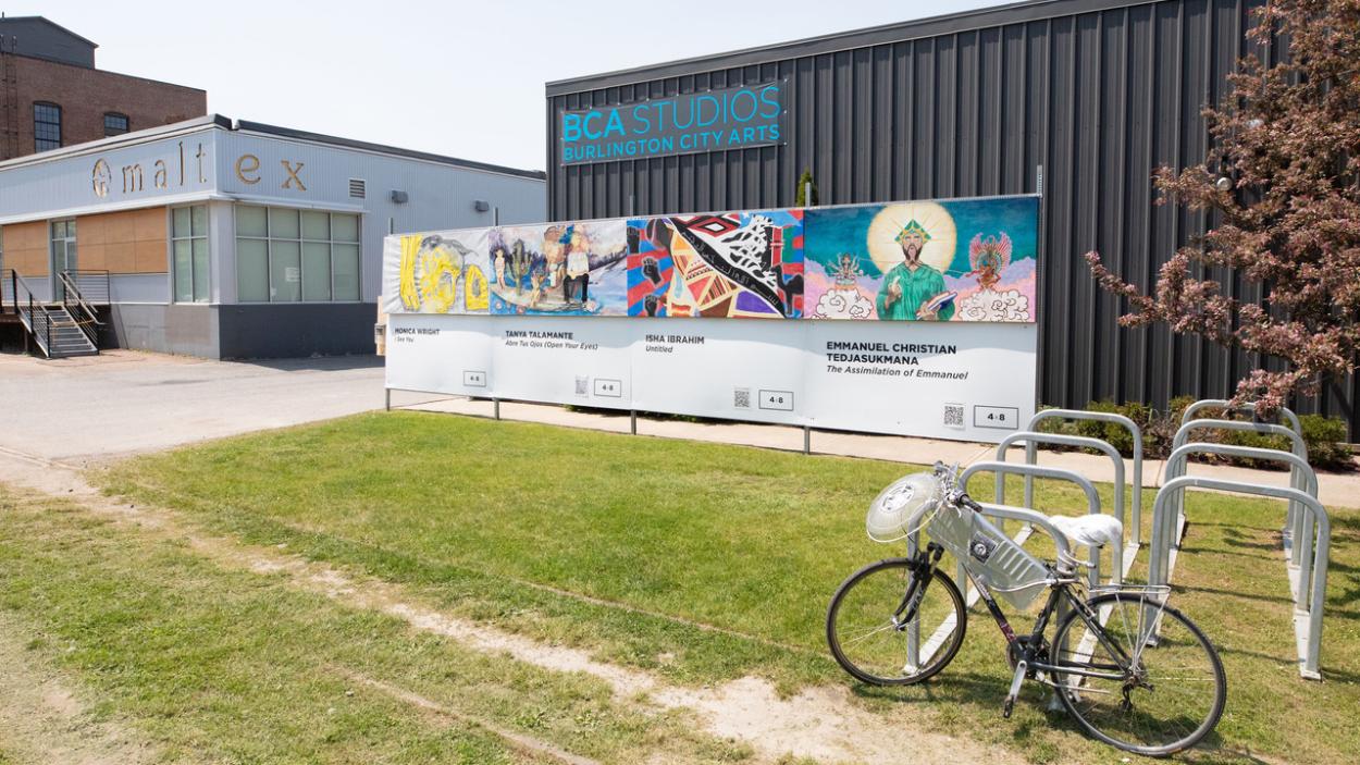 A photo of a square building with grey brown corrugated steel siding, and a sign that says BCA Studios: Burlington City Arts in blue lettering. A panel of 4 colorful murals, some grass and a bike rack with a bike in it sits in front of the building. A square blue grey building with a sign that says "Maltex" sits to the left.