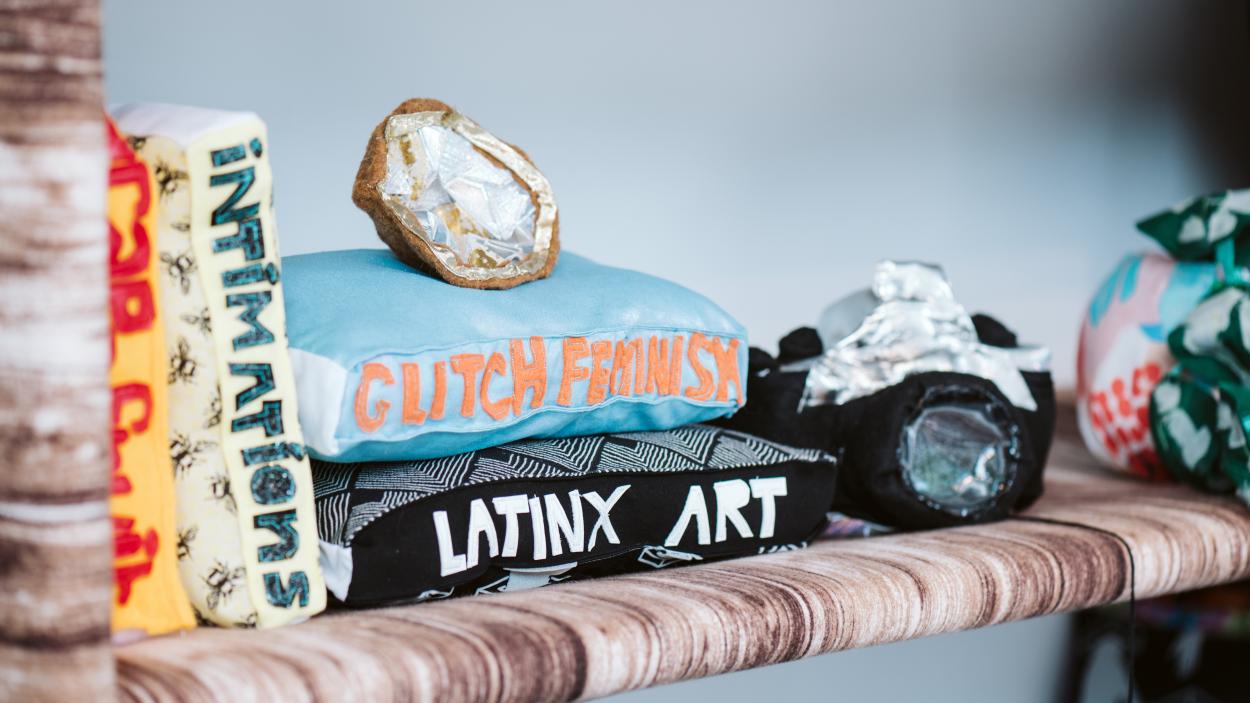 A photo of a detail of a soft sculpture, with books made out of textiles that say Intimations, Glitch Feminism, and Latinx Art. They sit on a shelf made from brown and white variegated fabric alongside a black camera made of fabric and a silver and grey geode made of fabric.