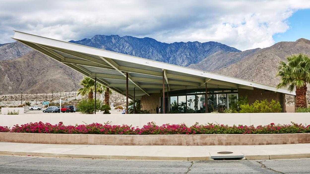 A photograph of a modern building with. white triangular dramatically slanted roof that overhangs a small glass building with brown and blue mountains and desert in the background.