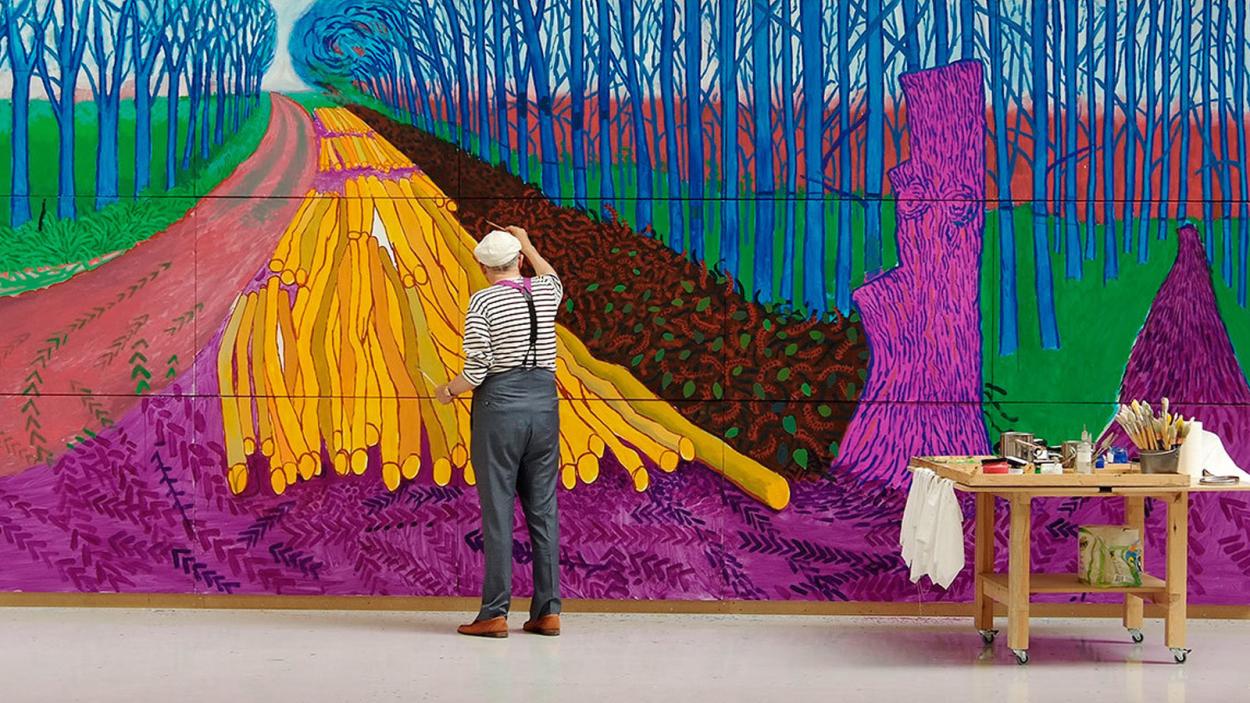 A photograph of David Hockney, a light skinned man with white hair wearing grey trousers and suspenders, working on a large painting with abstract shapes in fushia, green, saffron yellow, and teal.
