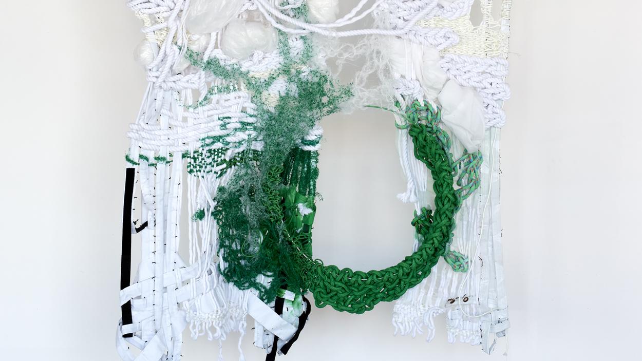 A textile sculpture that hangs on a white wall, consisting of knotted white cotton, plastic, and thread interwoven with rich green pieces that form a circle of negative space 