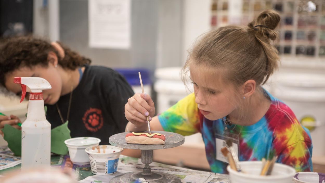 a light skinned girl, with dark blonde hair pulled back in a messy bun, wears a rainbow tie-dyed tee shirt and leans in intently as she paints a ceramic hot dog on a small pedestal. Clay tools, a spray bottle and another child wearing a black tee shirt are visible in the background 