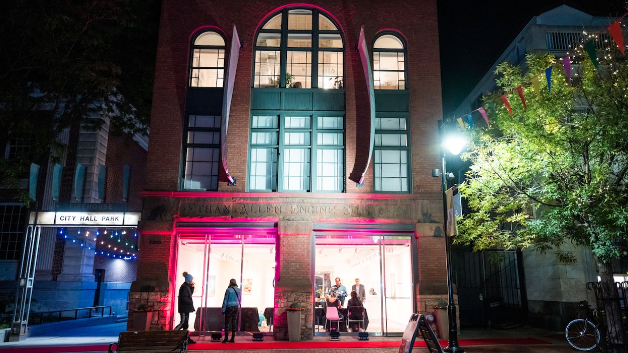A photo of the BCA Center at night. A narrow 4 story, red brick building with tall, arched windows, and aqua vertical banners, lit up inside, with people visible through the gallery doors