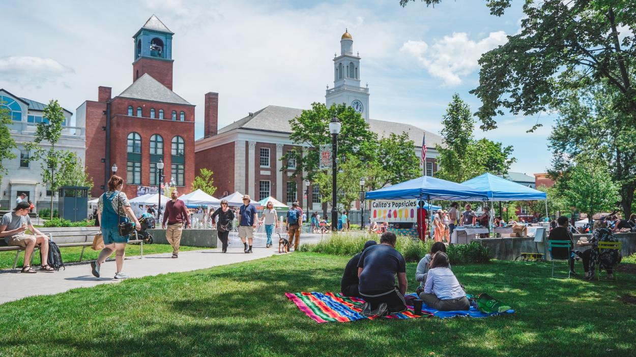 A photograph of Burlington's City Hall Park during the BTv Market. Vendors are set up under blue and white pop up tents, while people walk down the concrete paths, sit on blankets on the grass in the shade, and browse booths.  