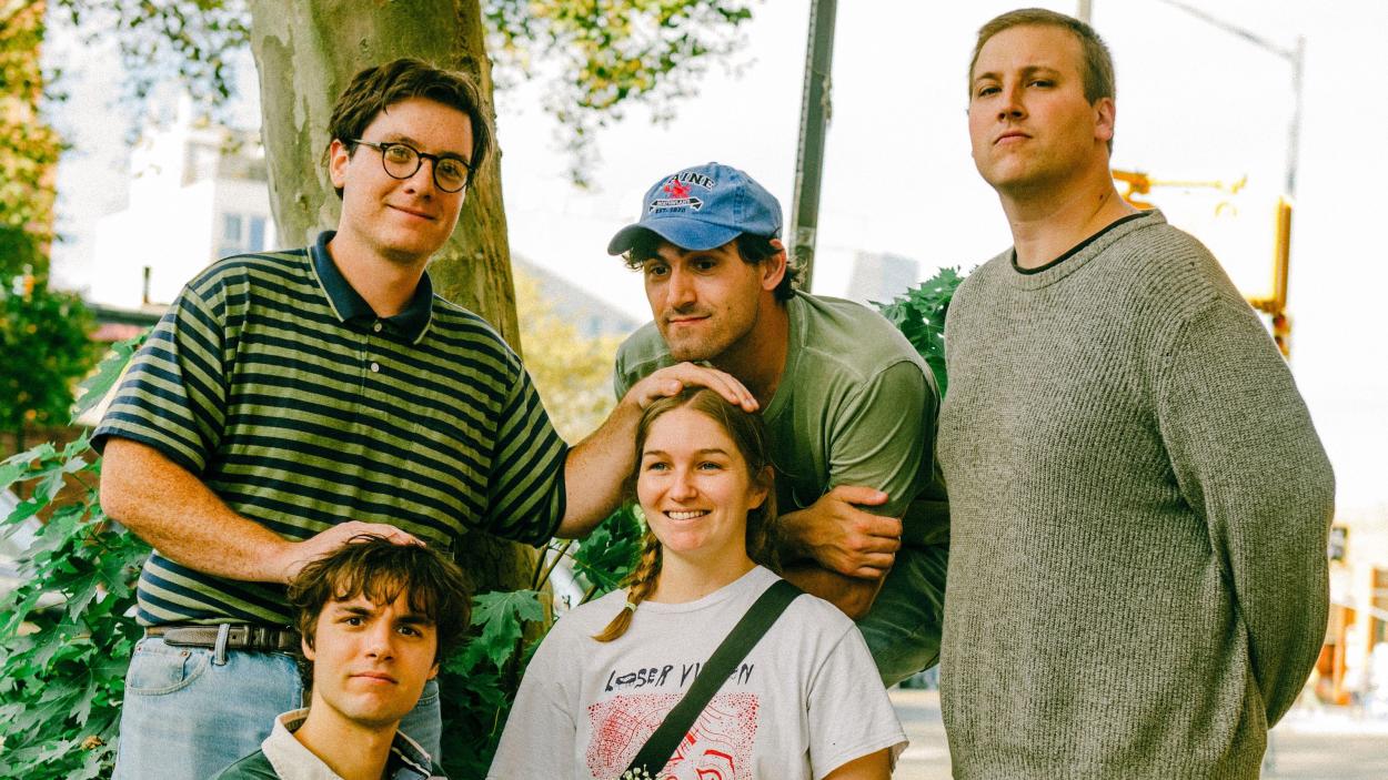 Four light skinned men and one woman are clustered together. One man wearing round glasses and green and black striped polo shirt stands on the left, with his hands on top of the head of a man who is sitting on the ground and the seated woman. Another man wearing a blue baseball cap leans over the seated woman and stand next to another man wearing a light green sweatshirt.