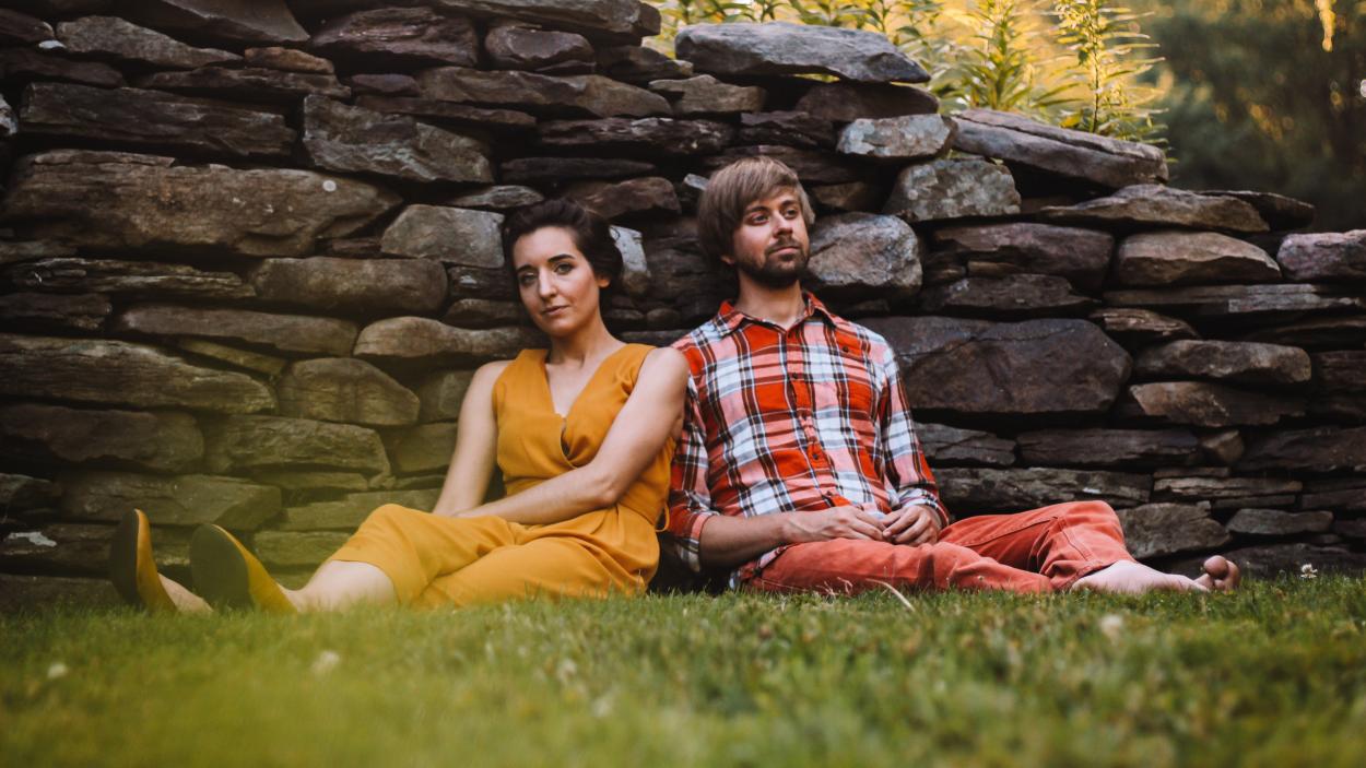 A man and a woman sit on the ground, leaning together against a stone wall. The woman has light skin and dark hair and wears a dark yellow  dress and shoes.The man has light skin and light brown hair and wears a red white and blue plaid shirt and red pants