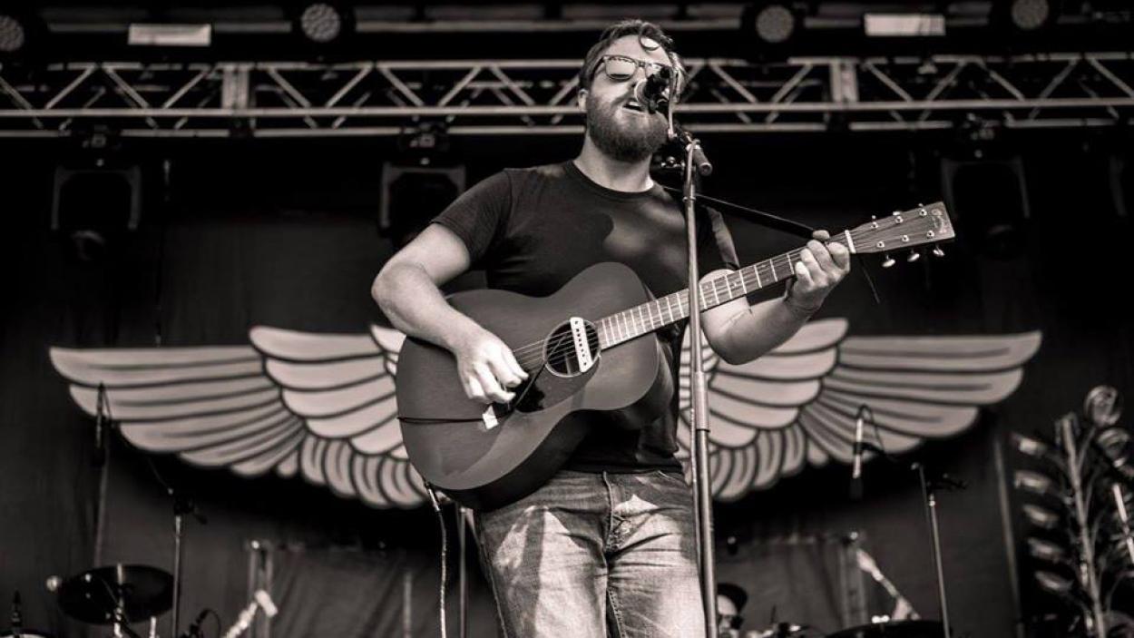 Eastern Mountain Time Promo.jpg A black and white photograph of a light skinned man with a dark beard and hair playing a guitar and singing into a microphone on a stage, framed by a decorative pair of wooden wings
