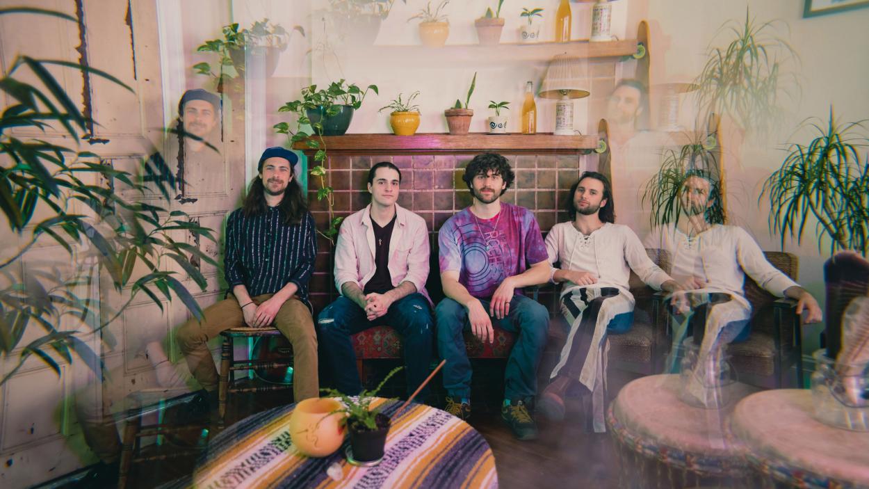 Head to the Roots (1).jpg Four young men, all with light skin and dark hair, sit in a row in a living room filled with house plants. The photograph has been double exposed so ghostly duplicates of parts of the image hover around the edges