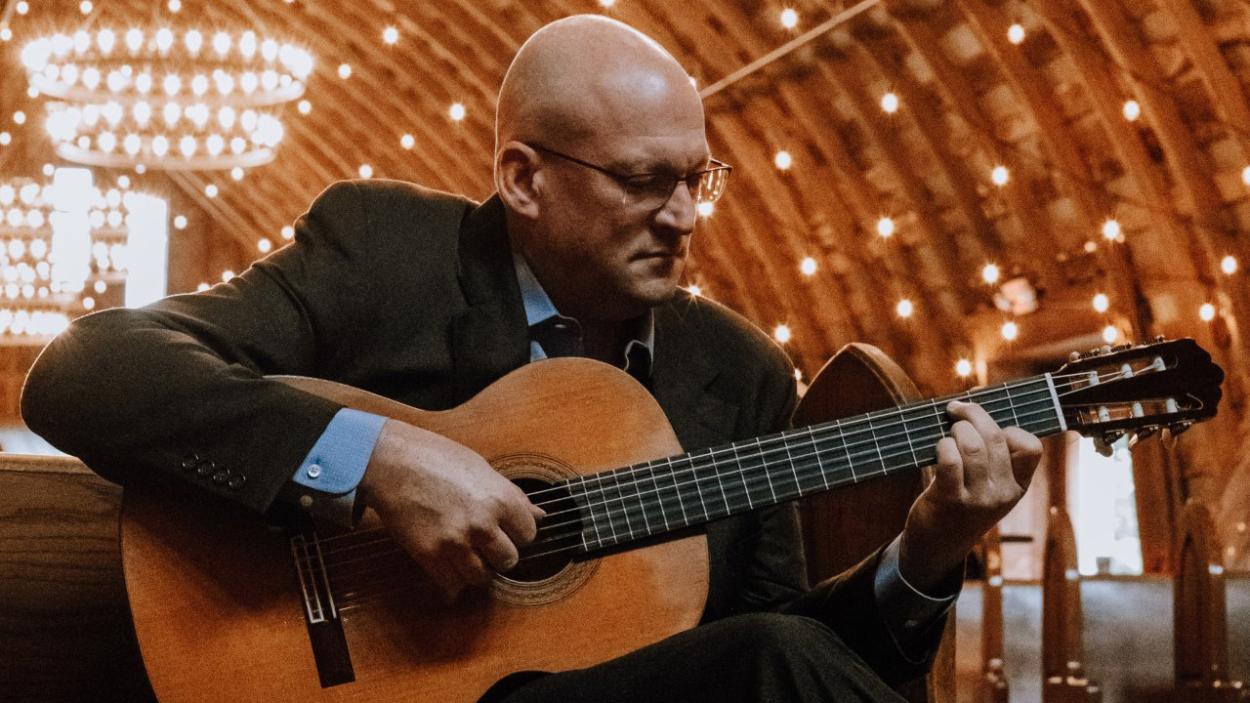 Kenneth Meyer 1.jpg A light skinned bald man wearing glasses and a dark suit plays an acoustic guitar. He sits in a pew under a wooden vaulted ceiling studded with points of light