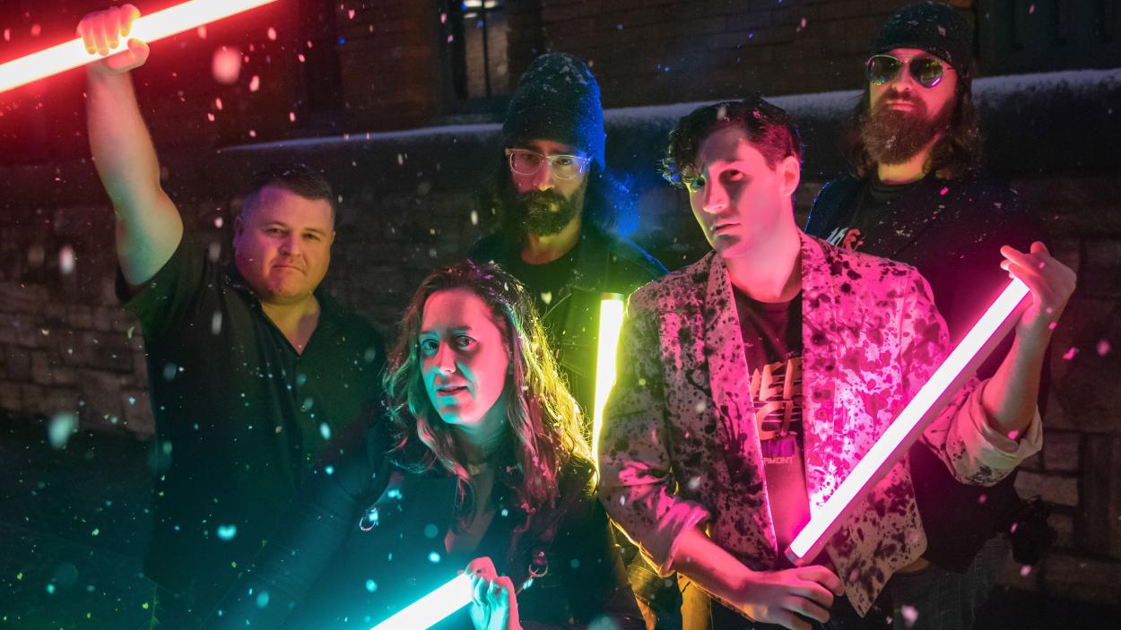 Night Protocol 2.jpeg Four light skinned men and one light skinned woman stand in the snow at night, illuminated by the colorful fluorescent tube lights they hold