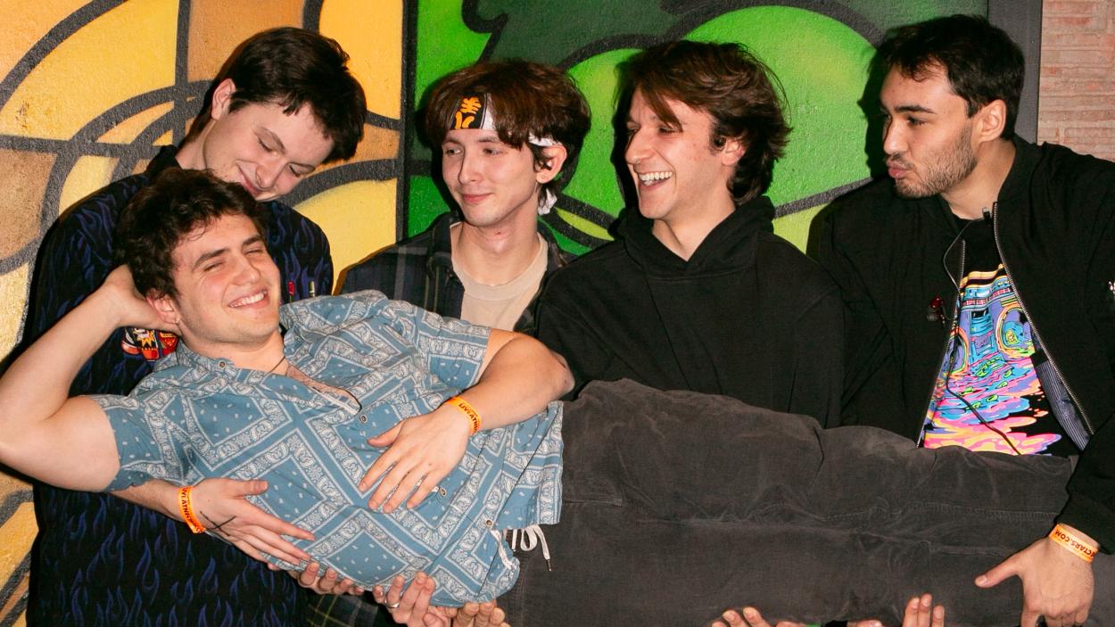 three young men with light skin tones and one with a medium skin tone laugh as they all hold another light skinned young man, laying horizontally