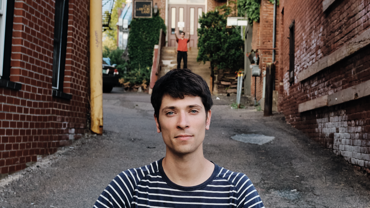 The Dead Shakers Press Photo by Ben Collins.png A light skinned man with dark hair and a black and white striped tee shirt is seen from the shoulders up. He stands in an alleyway, between brick buildings and in the background another man is positioned to look as is he is standing on the first man's head.