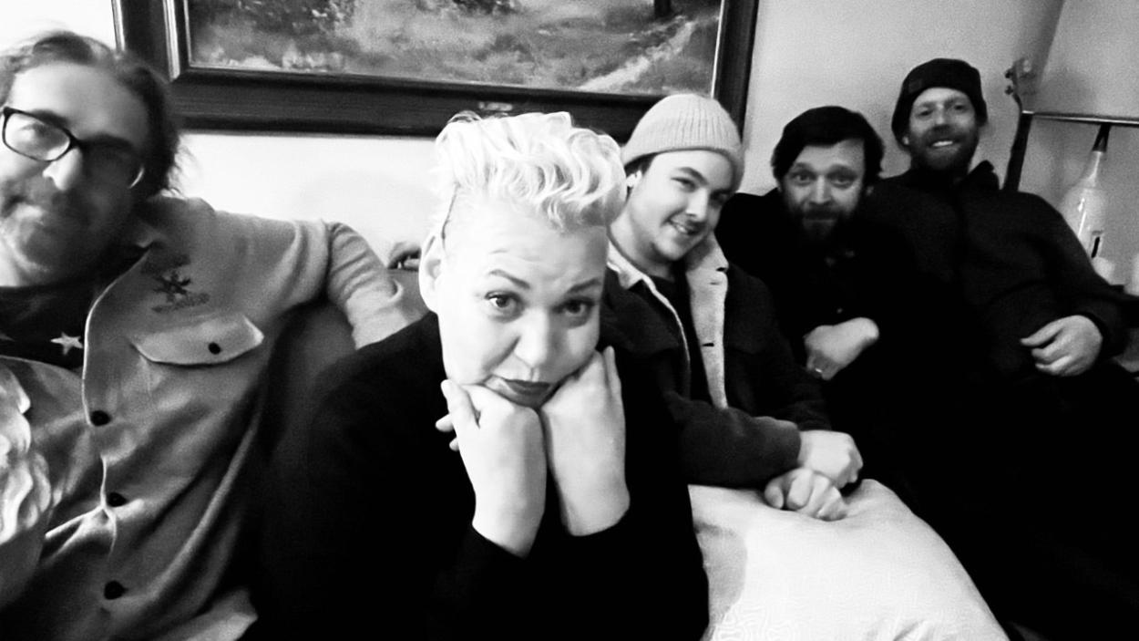 A black and white image of four light skinned men and one light skinned woman sitting on a couch. The woman who has a bleached blond pompadour leans forward and rests her chin on her hands