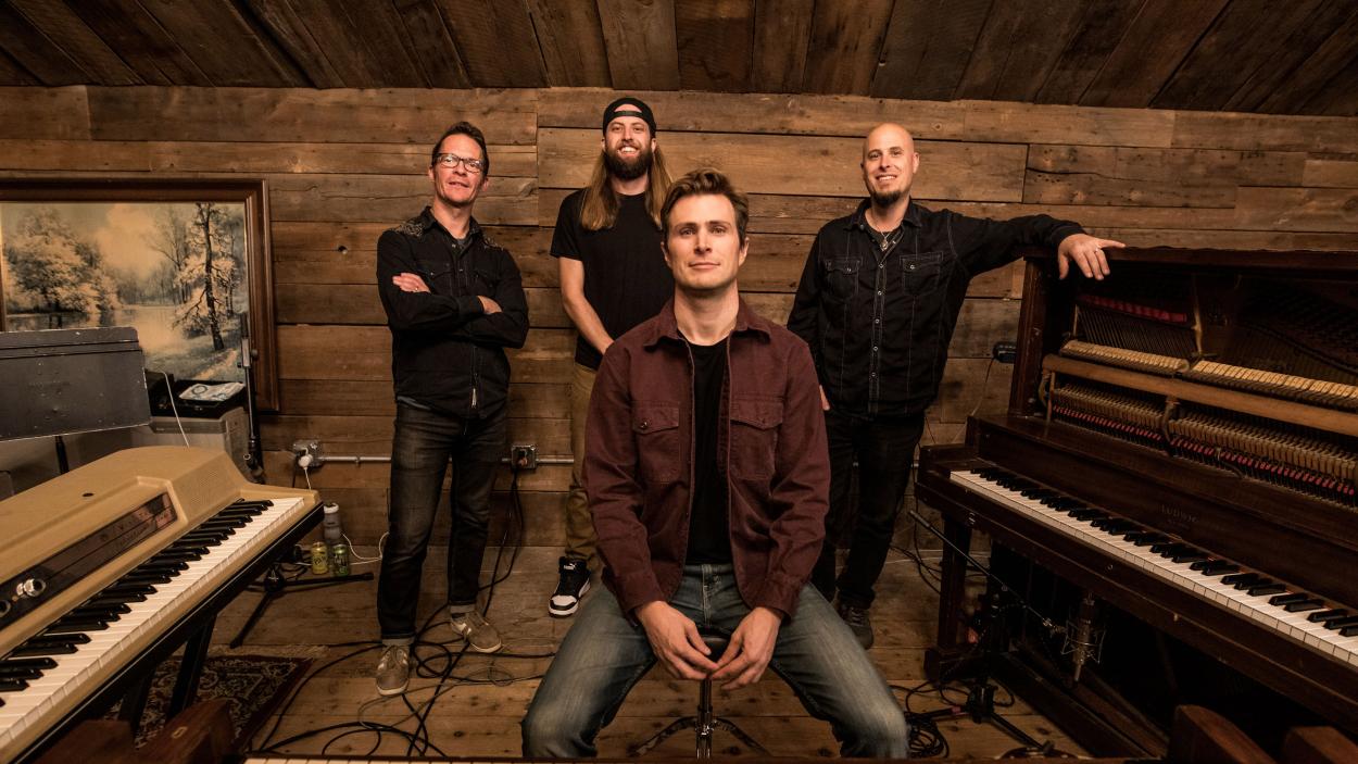Tyler Mast Full Band (hi res).jpg One light skinned man sits, while three ther stand behind him. A piano sits on one side and a keyboard on the other