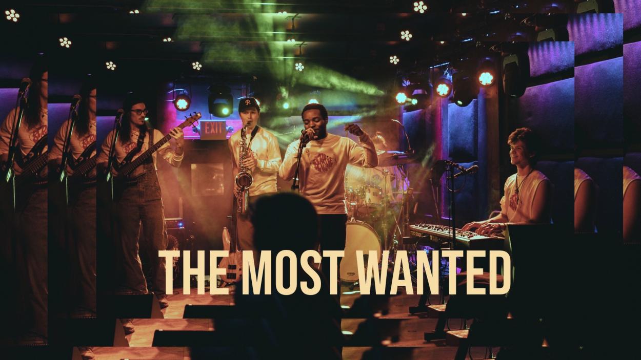 The Most Wanted over a picture of a band playing in a dark club, dimly lit with purple, blue and green lights. A dark skinned man wearing a white long sleeved tee shirt sings into a mic, while a light skinned man plays the saxophone behind him. On the left a light skinned man with long dark hair and glasses plays a guitar and on the right a light skinned man plays the keyboard. The image refracts, like a kaleidoscope at the outer edges.