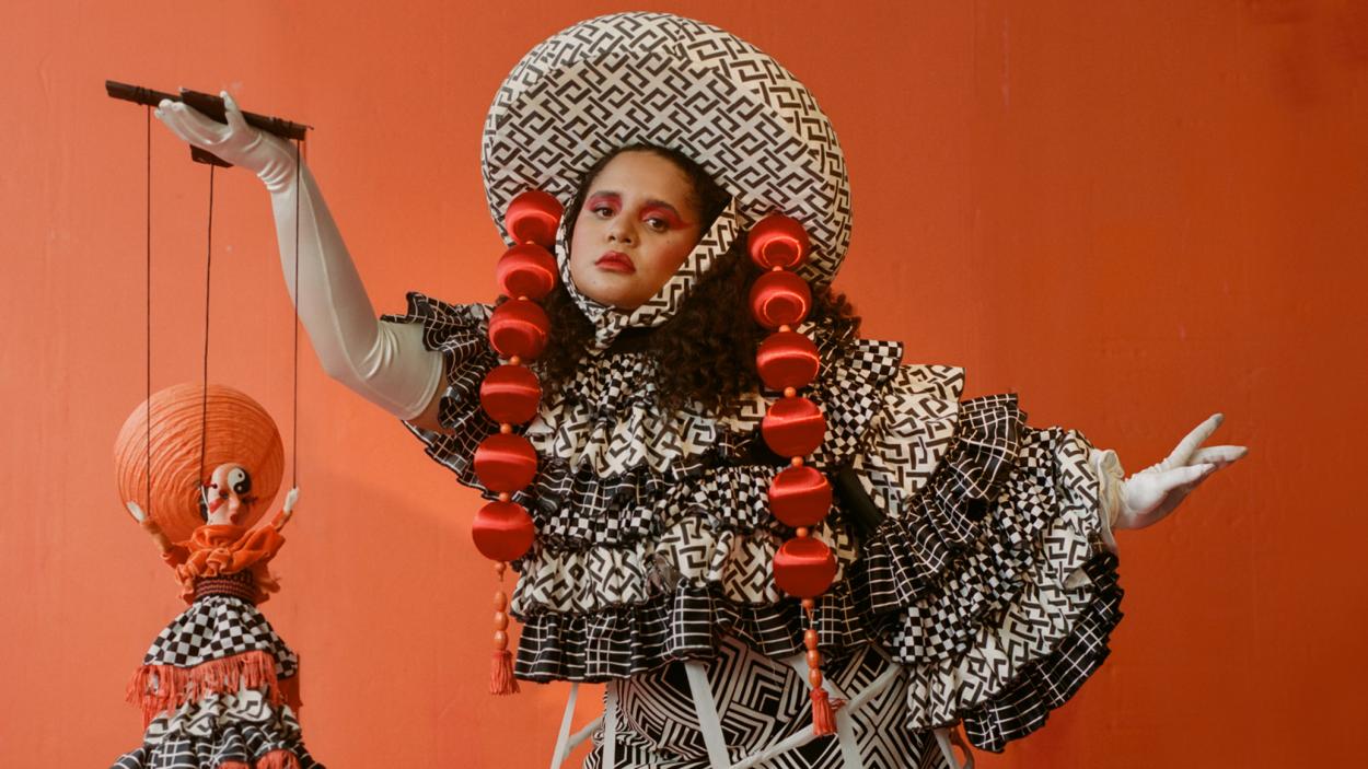 A medium skinned woman with dark hair wears an elaborate black and white ruffled patterned ruffled dress with a matching wide brimmed hat trimmed with two long strands of red balls that hang down beside her face. She stands against an orange backdrop and hold a marionette dressed in a similar style in one hand