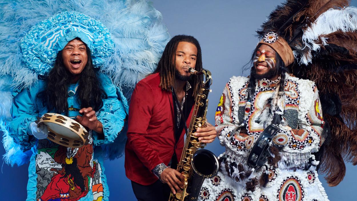 3_1.jpg Three dark skinned men play musical instruments against a light blue background. On the right, one man wears a large blue feathered headdress, a blue patterned and ruffled coat, and plays the tambourine and sings. In the middle, one man wears a red blazer and plays the saxophone. On the left one man has orange and yellow stripes painted across his cheeks and wears a large brown, black, and white feathered headdress and a white embroidered and patterned coat while he plays the tambourine