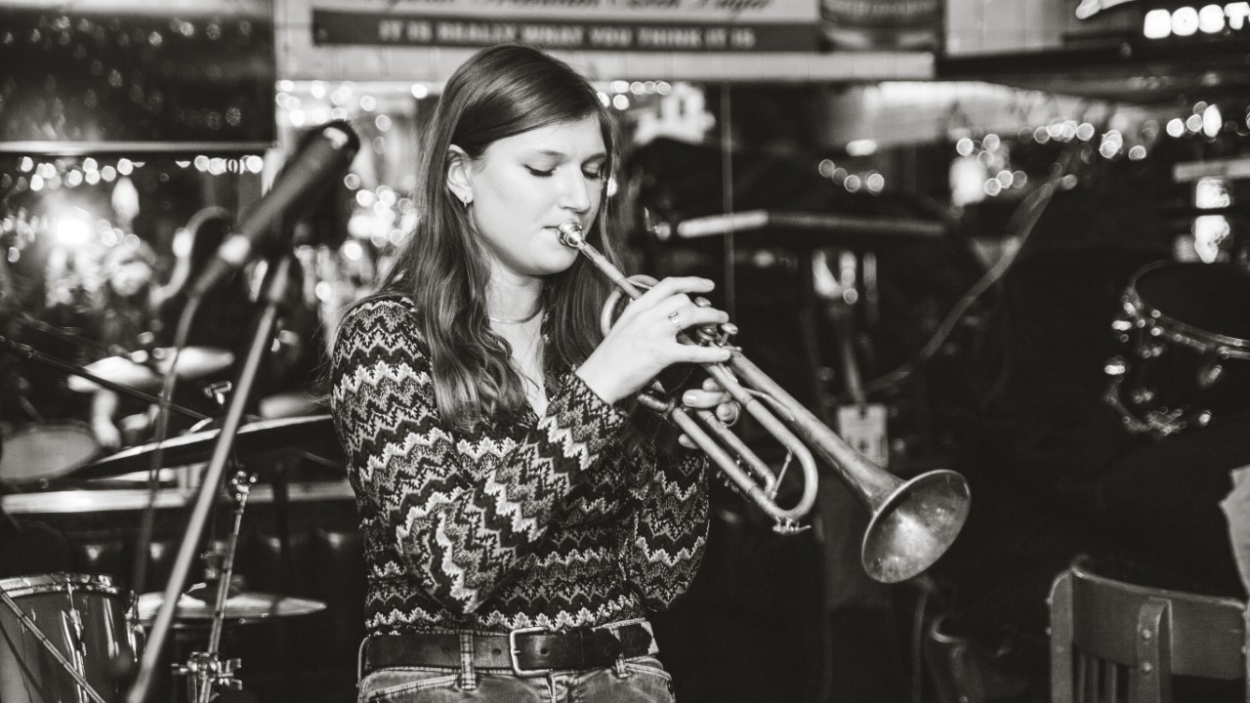clover_koval_3.png A black and white photograph of a young light skinned woman wearing a zig zag striped sweater and playing a trumpet in front of a microphone