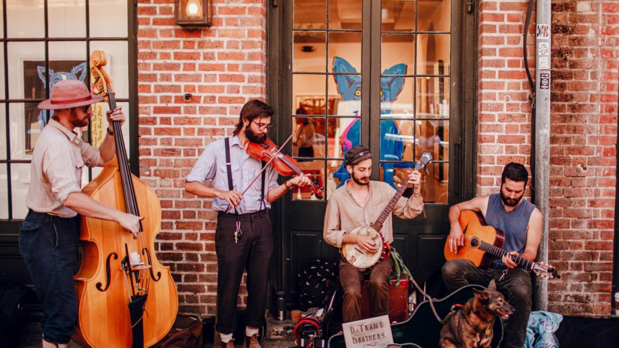 ditriani_brothers.jpeg Four light skinned men play musical instruments in front of a brick building. One wears a wide brimmed hat and plays a stand up bass, one wears a white shirt and black suspenders and plays the fiddle, one sits and plays the banjo, and one sits and plays an acoustic guitar