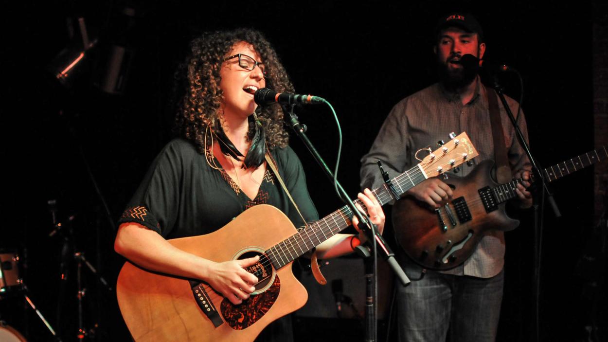 giovanina_bucci.jpg A light skinned woman with curly dark hair and dark rimmed glasses plays an acoustic guitar and sings into a microphone.