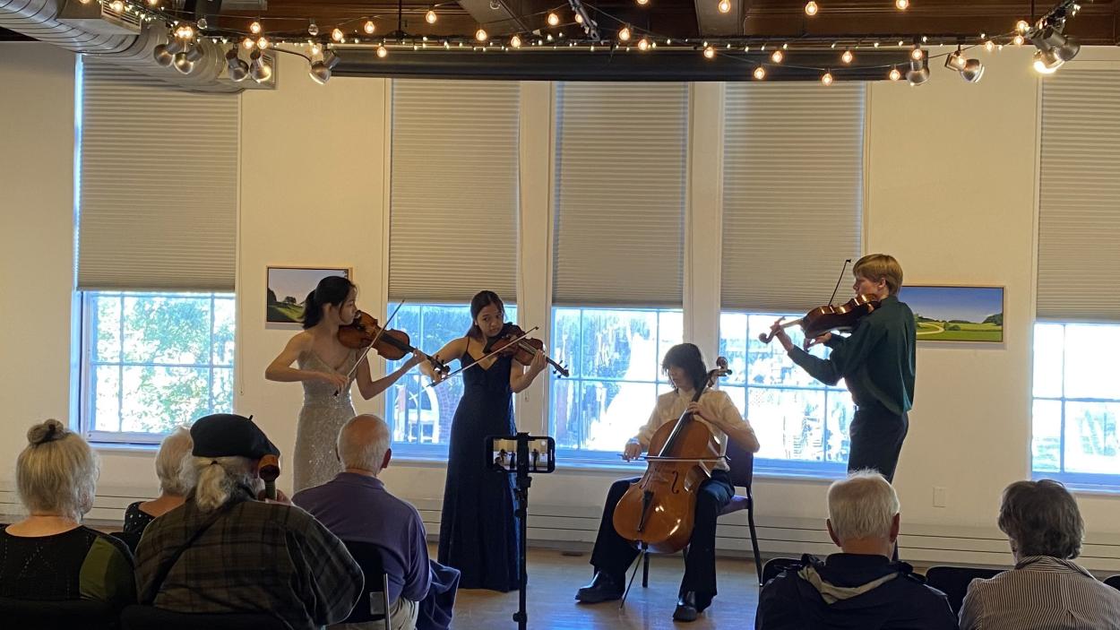 A quartet of young musicians play before an audience with tall windows with white shades half pulled behind them and twinkle lights above. Two young women play the violin, one wearing a long silver dress and one navy blue. One young man plays the cello and another plays the violin.
