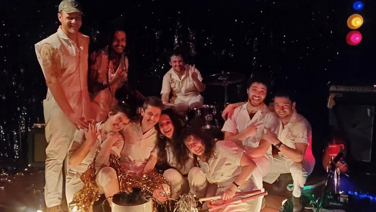 img_0334.jpeg Nine people sit, crouch, and stand clustered together. They all wear off white sleeveless jumpsuits. Some hold gold streamers. A sparkly curtain and stage lights are in the background