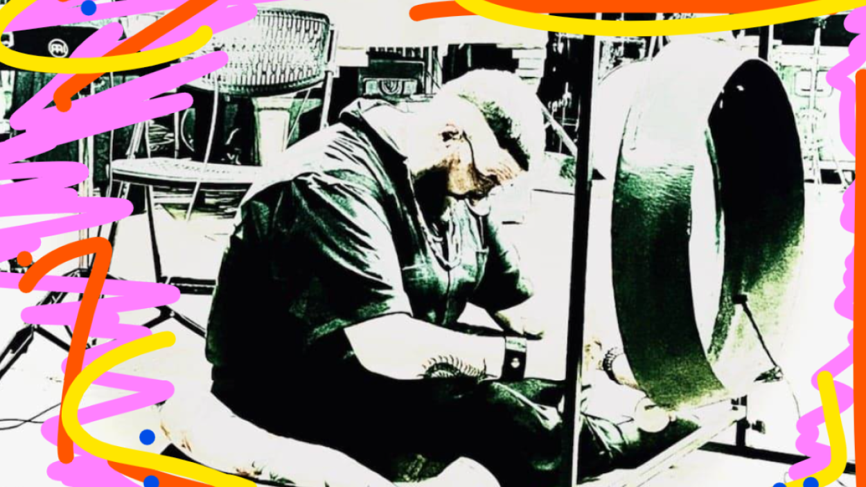 An image with a grey scale filter on it of a light skinned man with a beard wearing all black, sitting down and leaning forward. Neon pink and yellow zig zags are scribbled around the edges