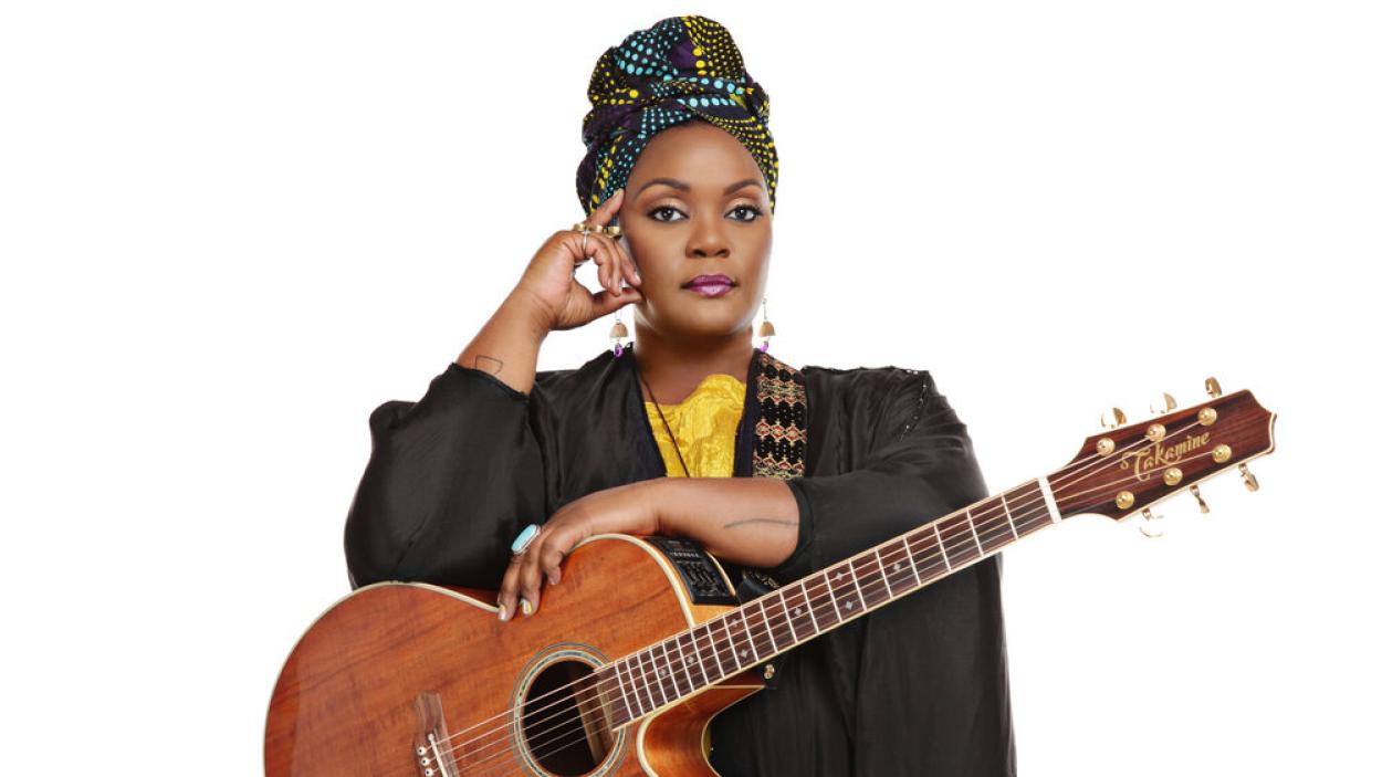 A dark skinned woman seen from the waist up against a white background. She wears a black jacket with a yellow shirt underneath, a black turban with a yellow and blue polka dot pattern and holds an acoustic guitar