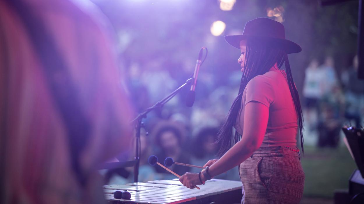 A dark skinned woman with long black braids wears a brown fedora and plays the vibraphone at an outdoor concert. She's seen from the side through a hazy, purple twilight and people sitting on the grass are visible in the background.