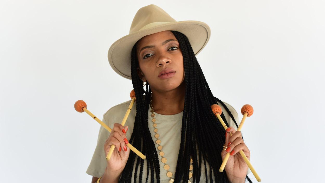 A dark skinned woman with long black braids wears a white fedora and top and holds four vibraphone sticks