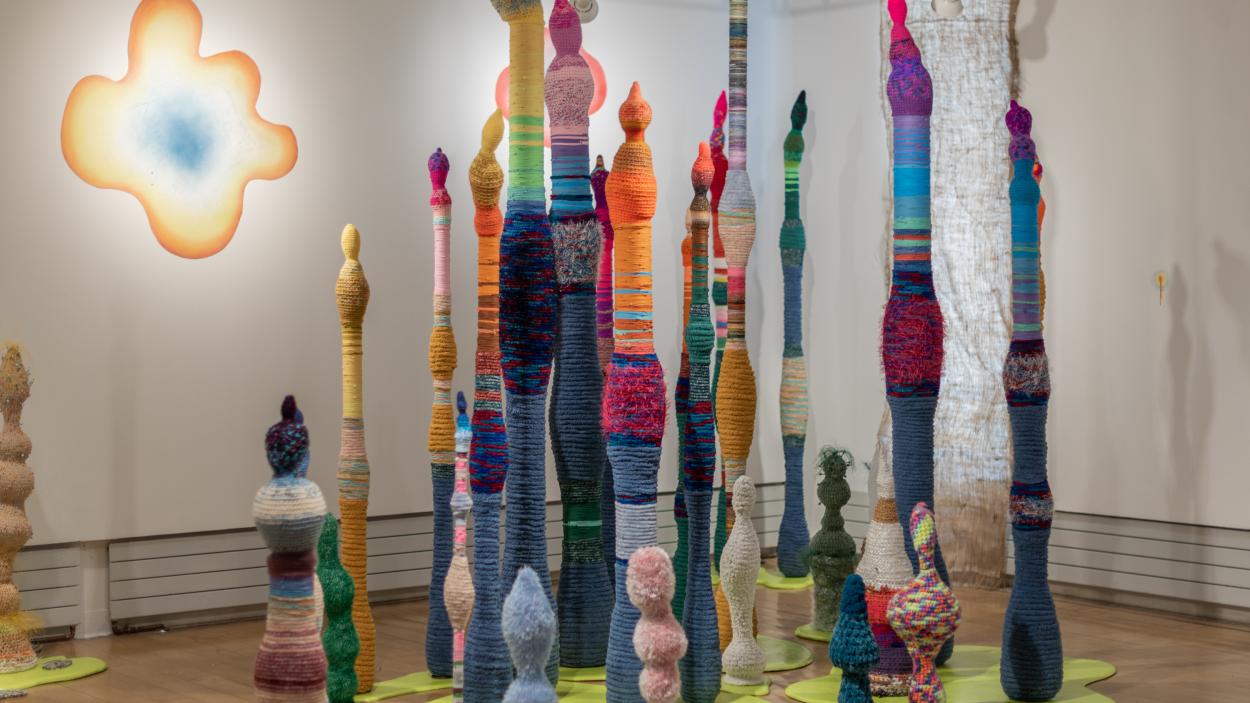 A gallery filled with tall, bulbous, linear multi colored sculptures with textured surfaces made from crocheted yarn. The sculptures rise from lime green bases in abstract blobby shapes. An abstract curvy blob that is blue in the center, fading to orange and yellow at the edges hangs on the wall in the background
