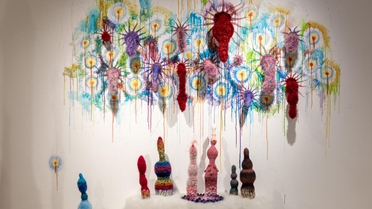 Bulbous, linear multi colored sculptures with textured surfaces made from crocheted yarn sprout from a wall, surrounded by multi-colored painted starbursts, while several other sculptures stand below on a white fuzzy mound. 