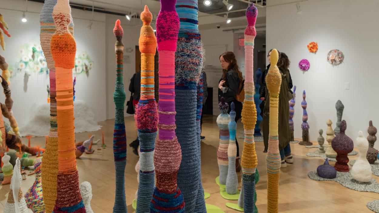 A gallery filled with tall, bulbous, linear multi colored sculptures with textured surfaces made from crocheted yarn. Several people mill around in the background
