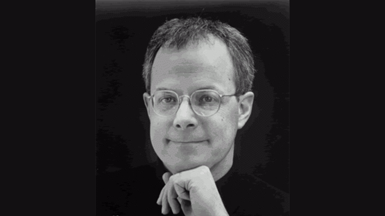 A head shot of a light skinned man wearing round wire rimmed glasses and resting his chin on his hand against a black backdrop