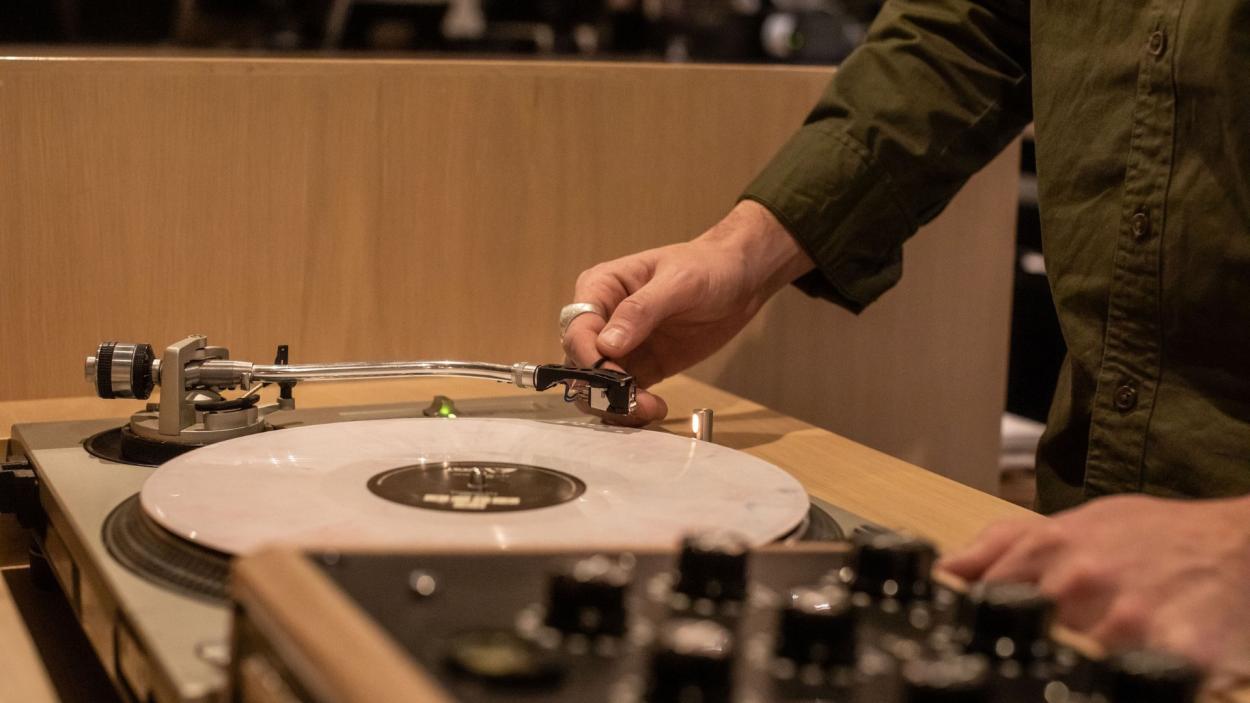 A cream colored record spins on an elaborate turntable set up, and the hands of a light skinned man wearing an olive green shirt can be seen lifting the needle. The back of a blonde wood banquette blond wood piece is in the background