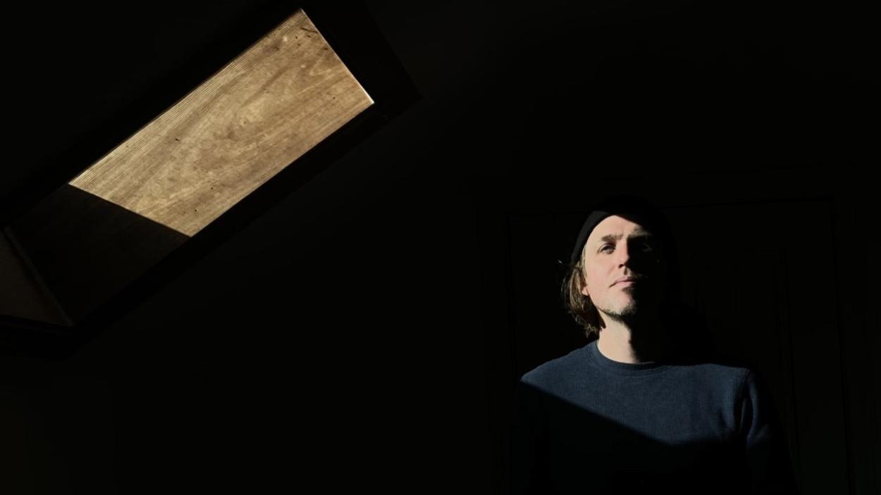 A light skinned man with light brown hair seen from the shoulders up, with his face half obscured by a dark background, interrupted only by a geometric wooden form floating above him