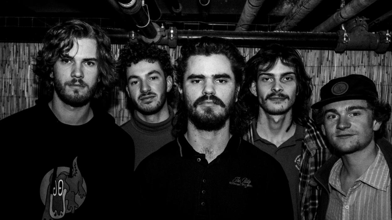 A black and white photo of 5 light skinned men seen from the chest up standing closely grouped together