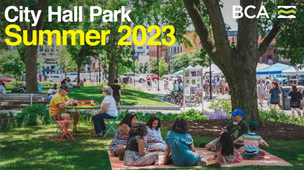 City Hall Park Summer 2023 BCA. A group of people sit on an orange blanket on the grass in the shade of a tree. Other people sit at colorful cafe tables near by and a street lined with vendor booths is visible in the background