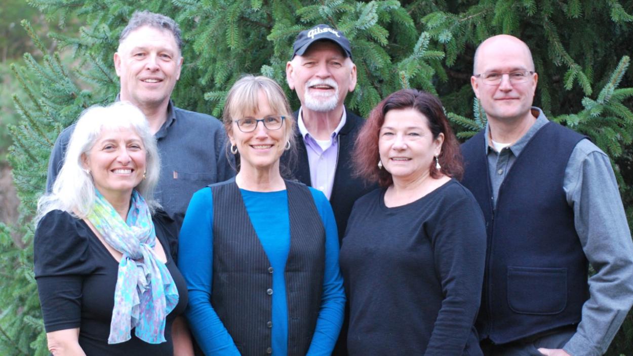 Three light skinned older men stand behind three light skinned older women, with leafy trees in the background